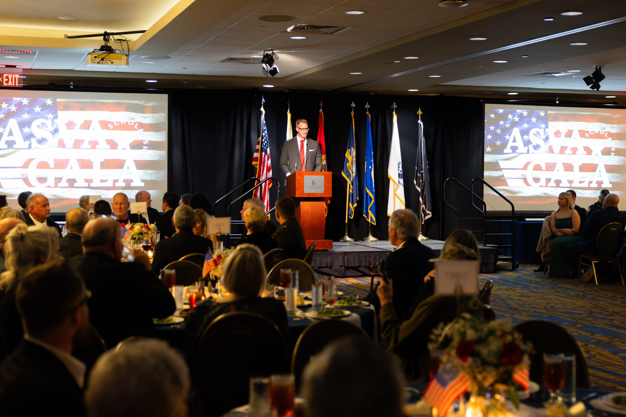 Man standing in front of a podium on a small stage speaking to a crowd of people seated at tables.